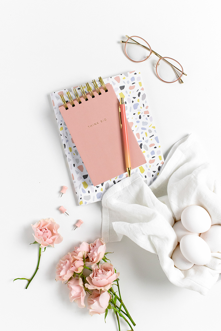 Flatlay of stacked notebooks with glasses and pink roses surrounding it